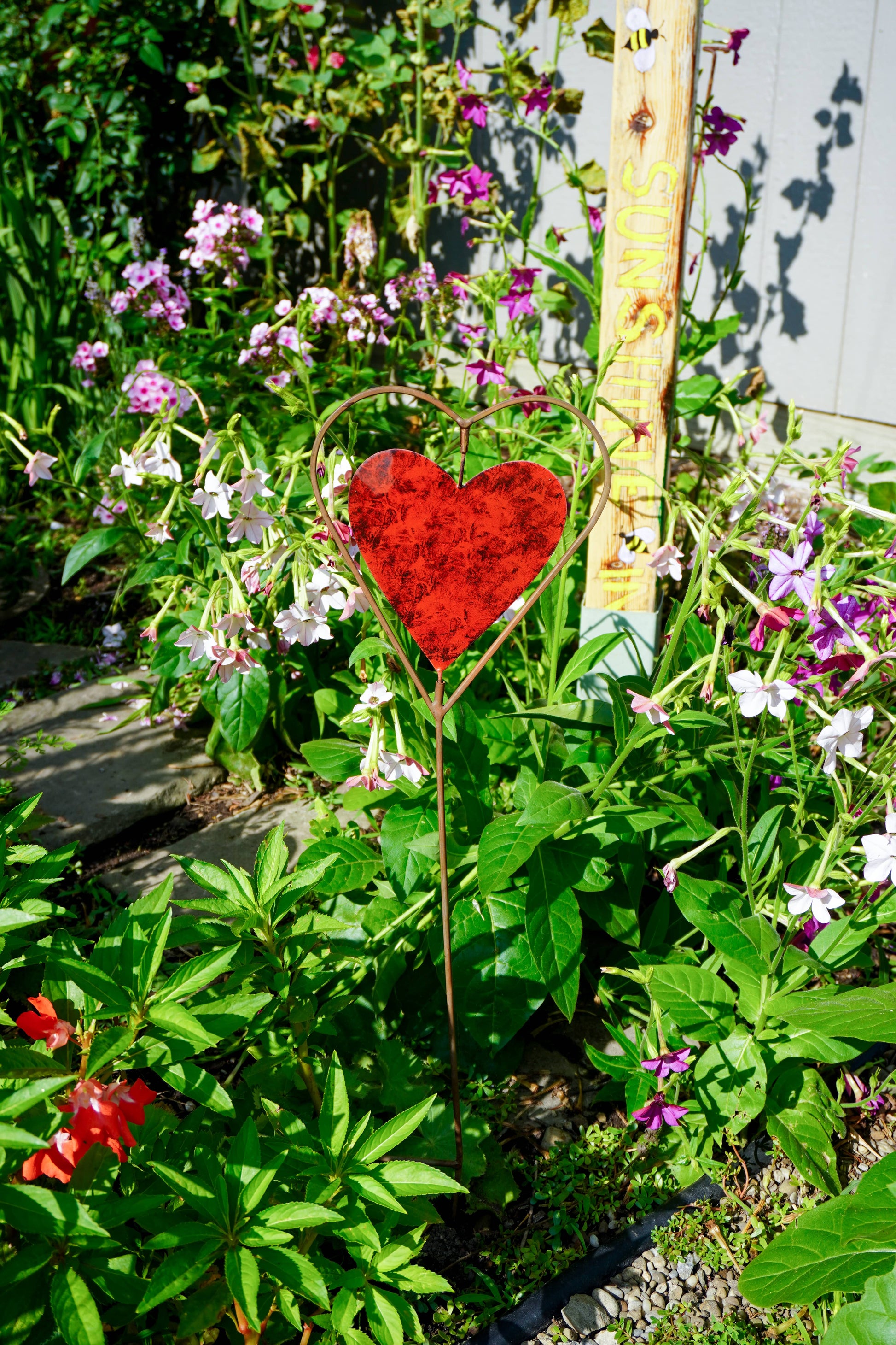 A red heart-shaped metal garden stake with a heart outline at the top, placed in a lush garden bed surrounded by green foliage and small pink and purple flowers.