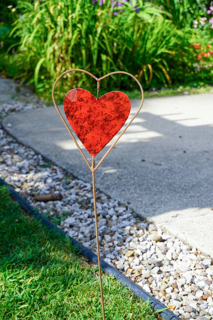 A decorative metal heart garden stake with a textured red heart center, placed in a grassy garden bed next to a concrete path.