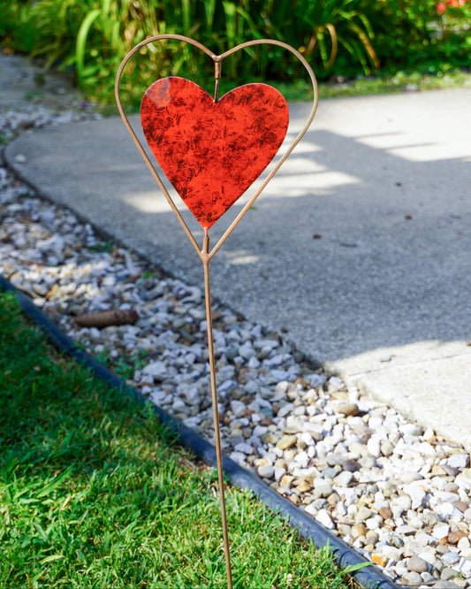 A decorative metal heart garden stake with a textured red heart center, placed in a grassy garden bed next to a concrete path.