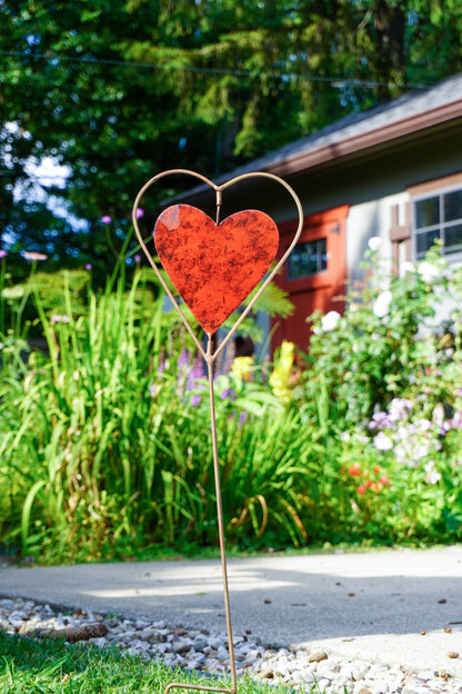 A red heart-shaped garden stake with a gold metal wire heart outline, placed in a green garden pathway in front of a house.