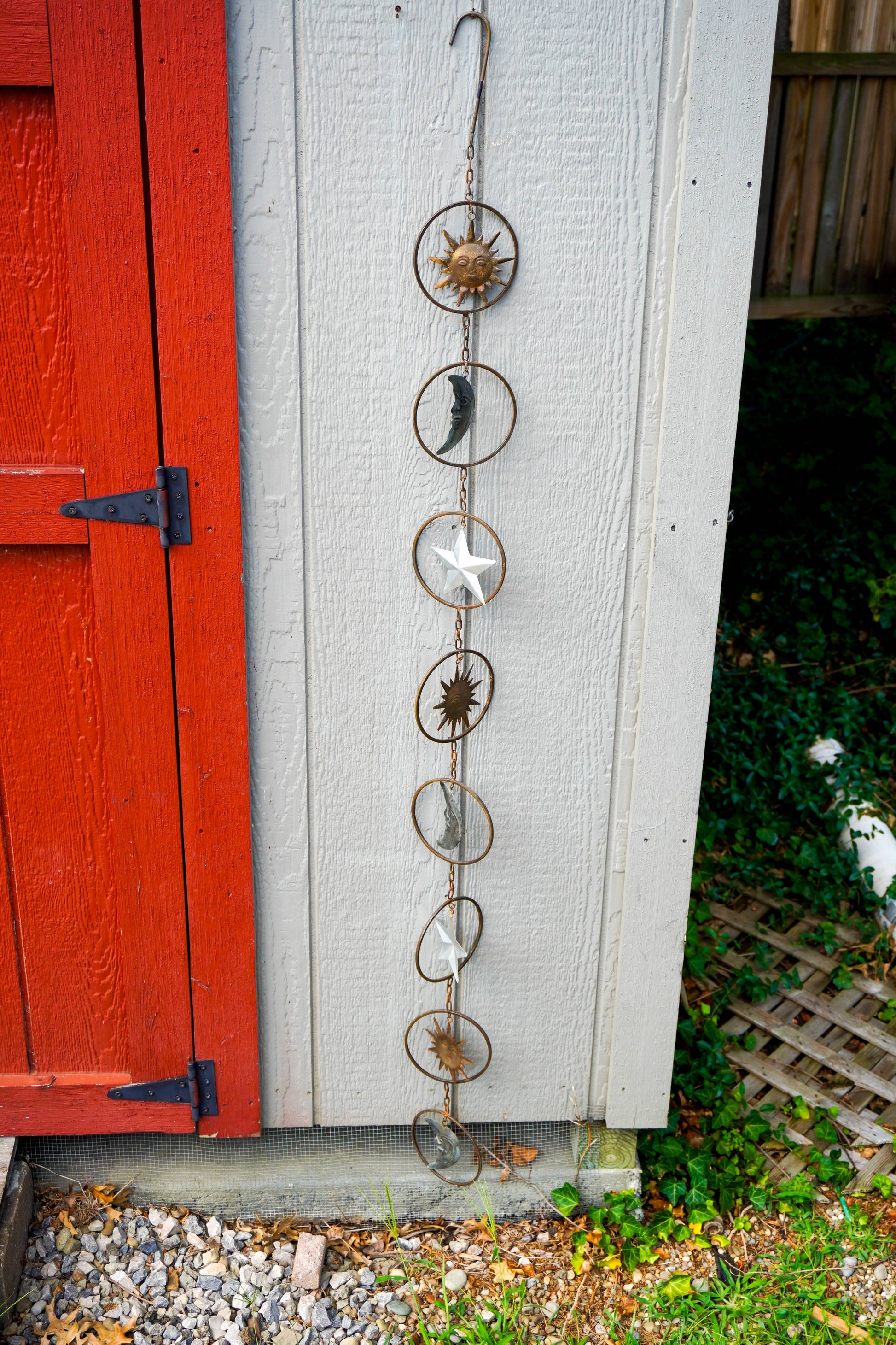A vertical metal moon phase hanging ornament with star and sun motifs, displayed outdoors against a white shed wall next to a red wooden door.