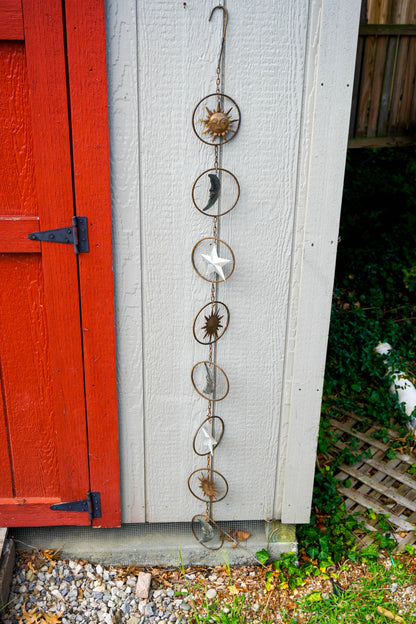 A vertical metal moon phase hanging ornament with star and sun motifs, displayed outdoors against a white shed wall next to a red wooden door.