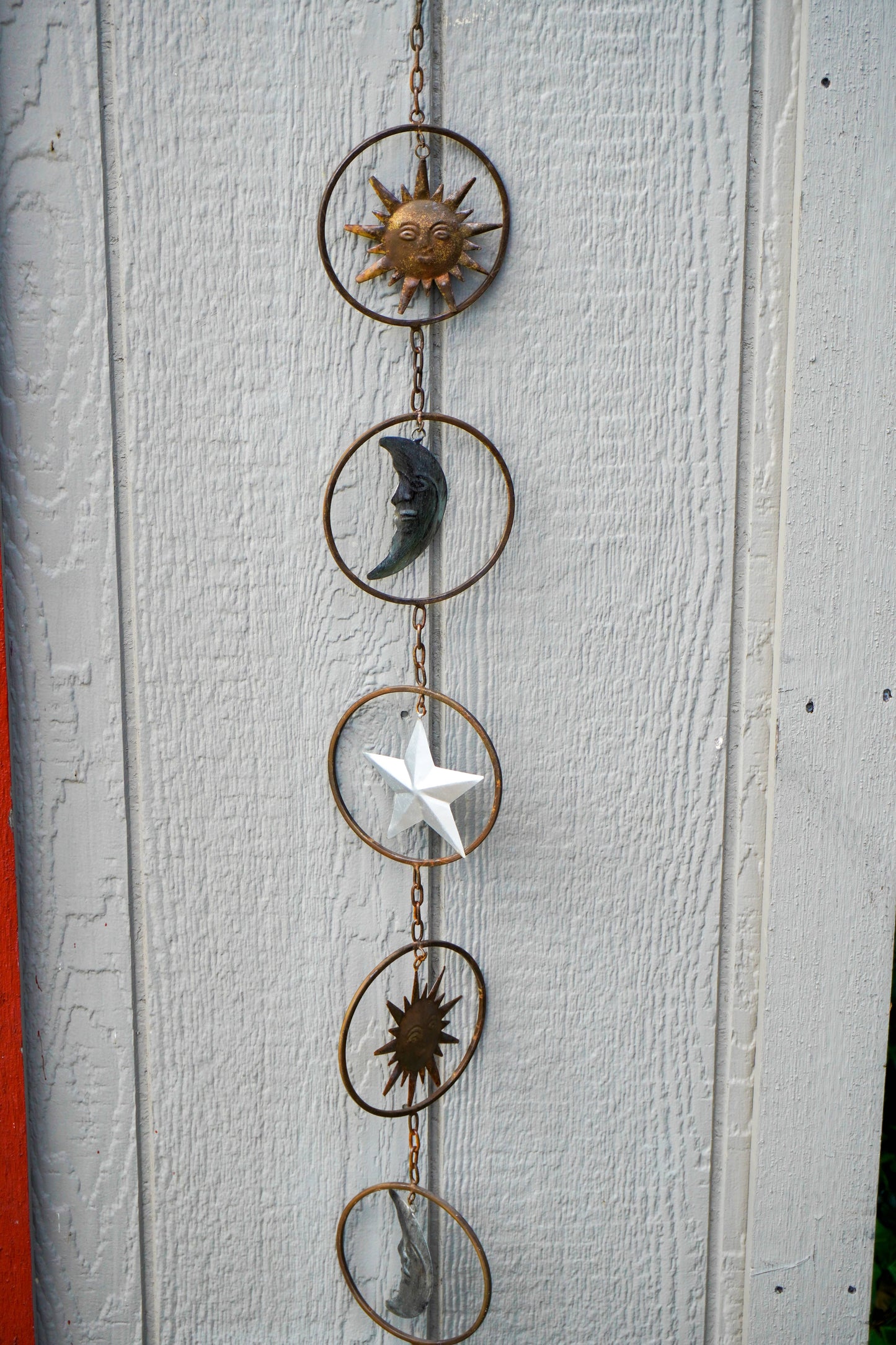 A vertical metal celestial hanging ornament with five circular rings connected by a chain, featuring sun, moon, star, and abstract sun motifs in different colors including gold, dark gray, white, and bronze, displayed against a white painted wood wall.