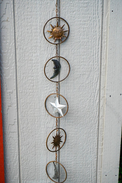A vertical metal celestial hanging ornament with five circular rings connected by a chain, featuring sun, moon, star, and abstract sun motifs in different colors including gold, dark gray, white, and bronze, displayed against a white painted wood wall.