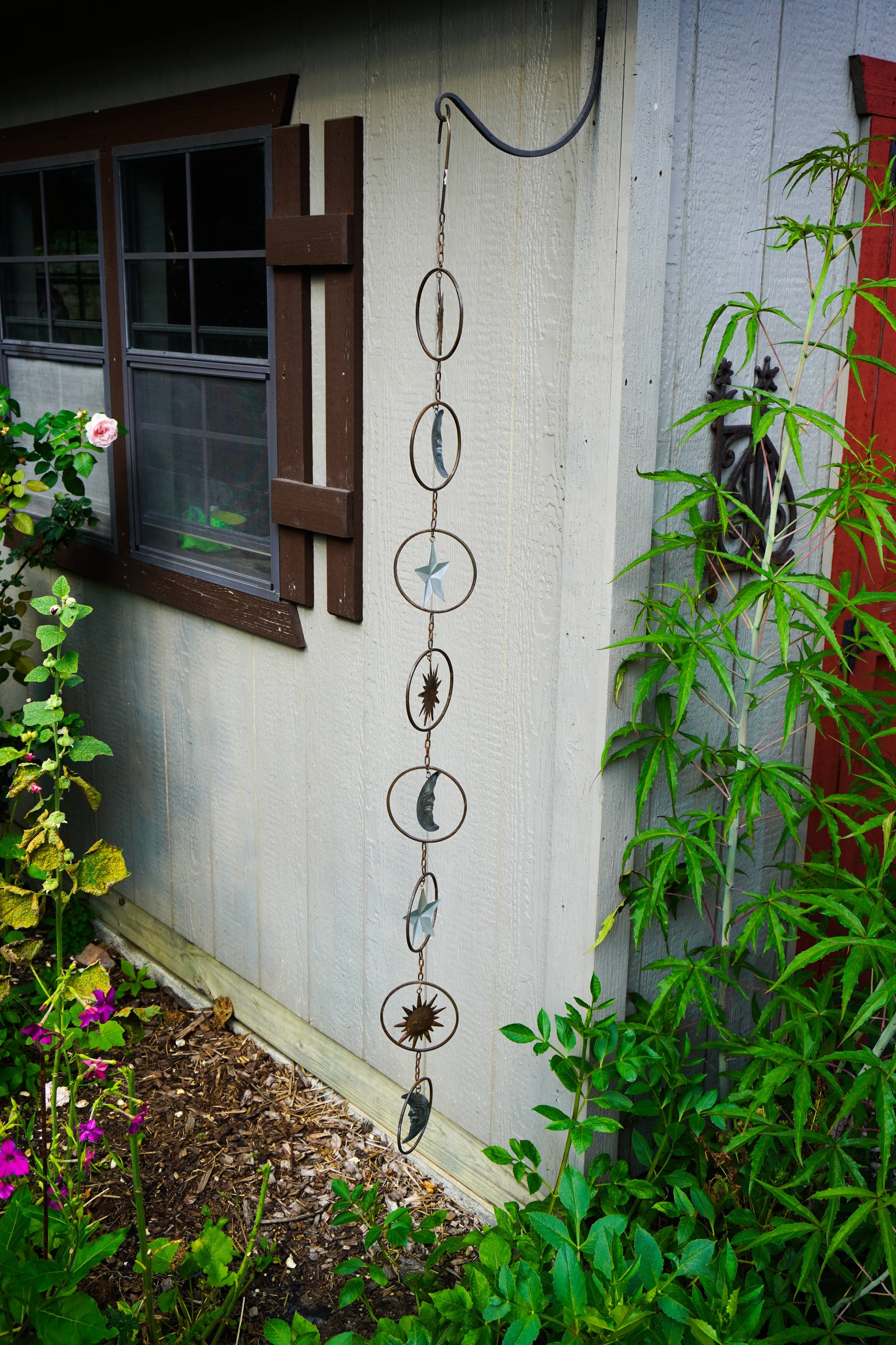 A hanging metal garden wind spinner with circular rings and moon phases, displayed outdoors on a white house wall next to green plants and a window.