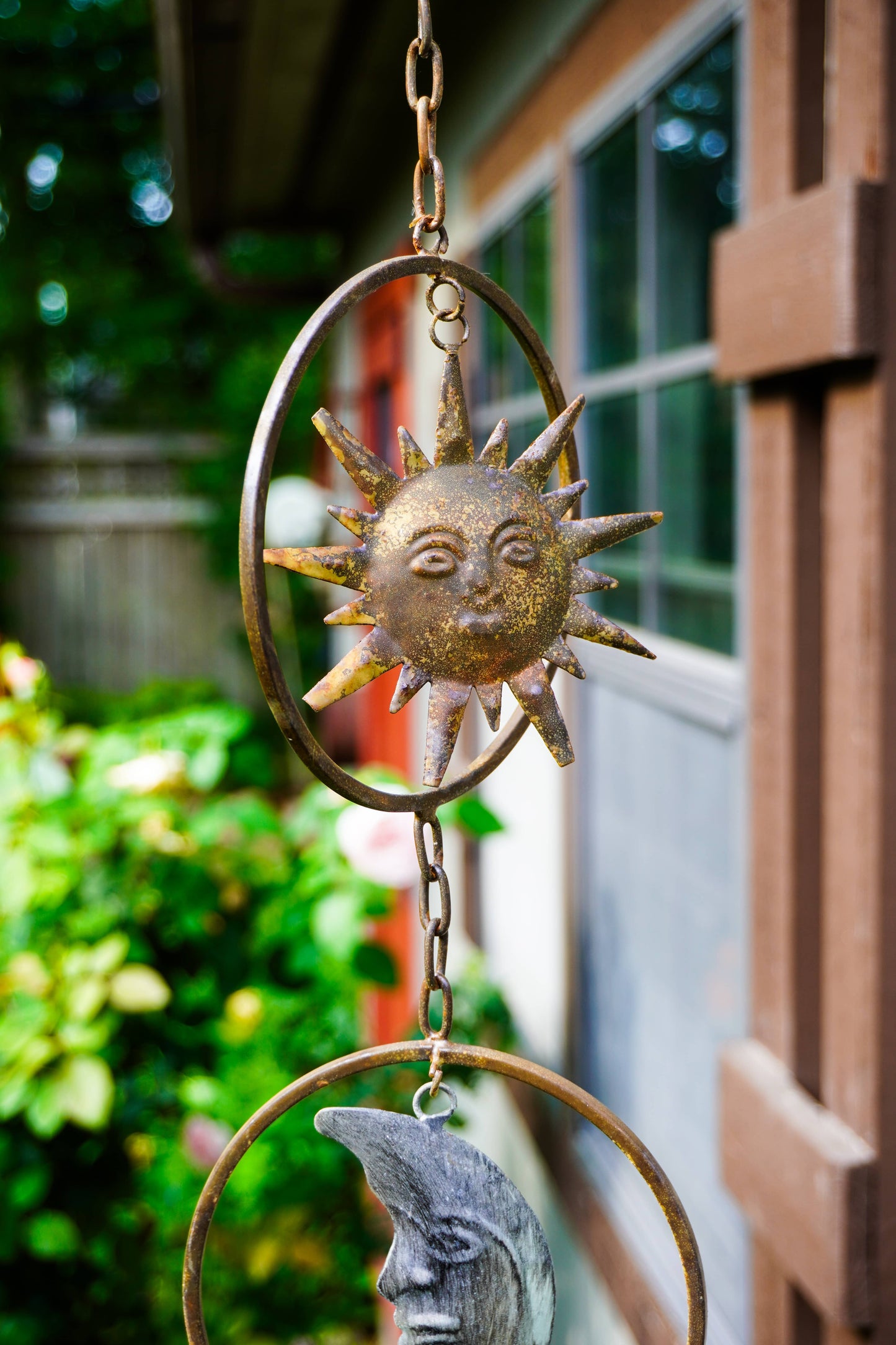 A hanging metal garden ornament featuring a sun face inside an oval frame, with a chain leading to a crescent moon face below, photographed outdoors near a wooden building and greenery.