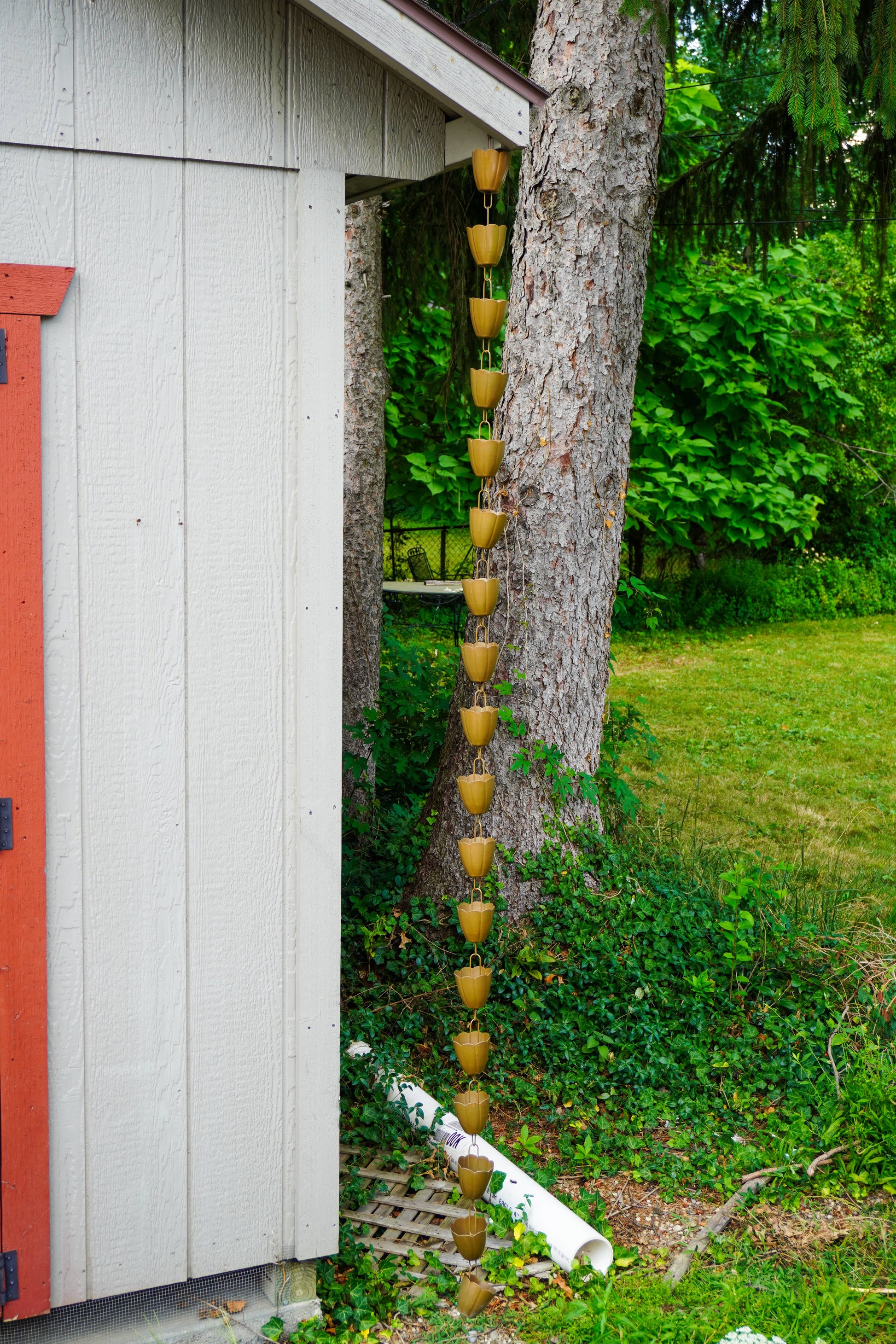 A vertical rain gutter hanging from the corner of a white-sided house next to a tree trunk, with a long chain of small metal pots suspended down the gutter in a garden setting.