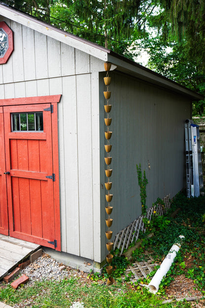 A vertical hanging rain gutter with multiple small gold-toned cup sections, installed along the corner of a gray shed with a red door in an outdoor garden setting.