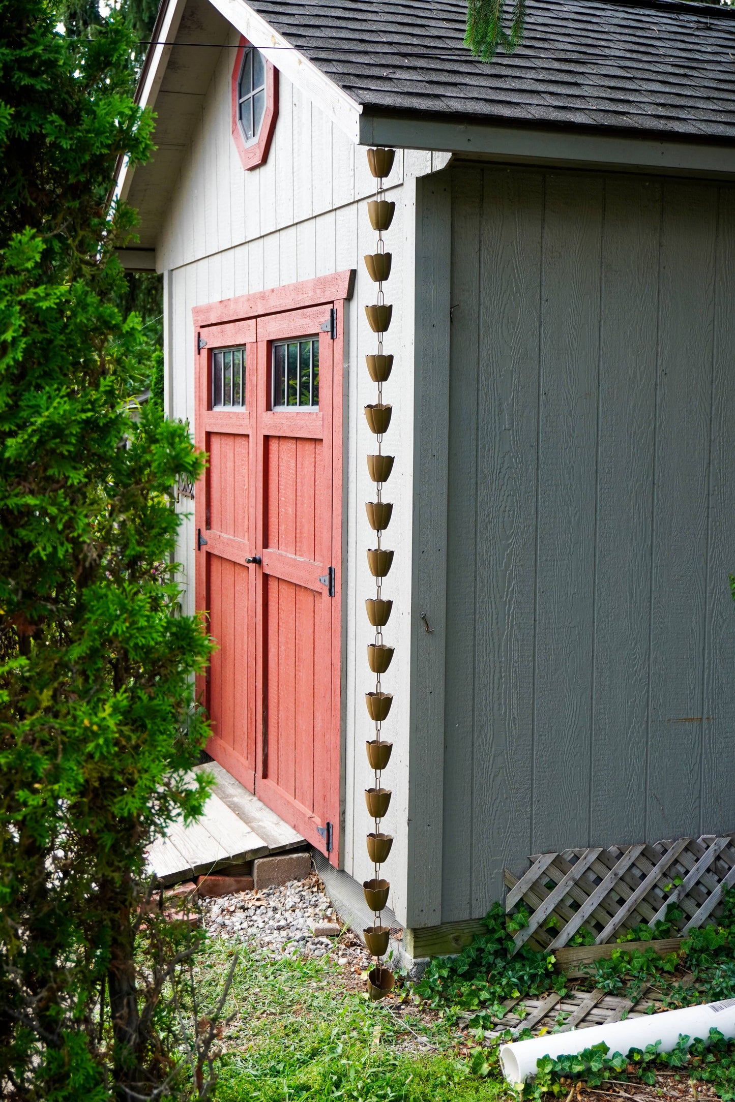 A rustic outdoor scene showing the corner of a small shed or cabin with a red double door, gray siding, a dark roof, and a hanging string of small brown pots or planters along the edge. Greenery and gravel are visible around the structure.