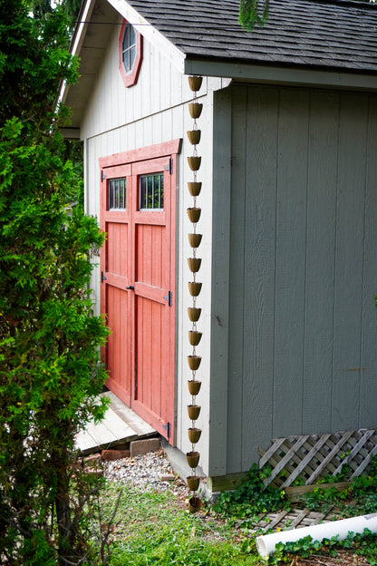 A rustic outdoor scene showing the corner of a small shed or cabin with a red double door, gray siding, a dark roof, and a hanging string of small brown pots or planters along the edge. Greenery and gravel are visible around the structure.