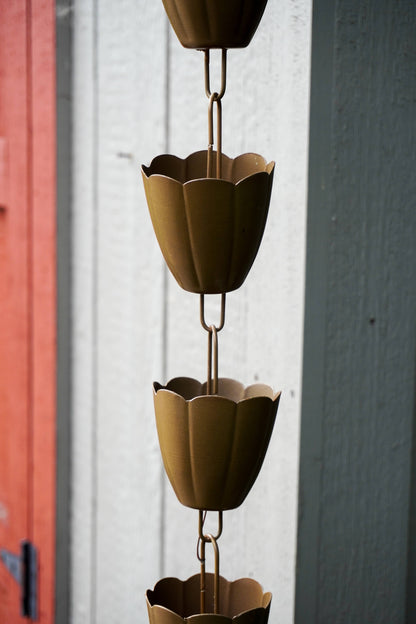 A close-up of a metal rain chain hanging vertically outdoors, with flower-shaped cup links in a warm metallic gold finish against a white wall and a red door frame.