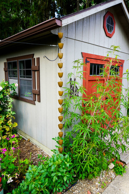 A vertical hanging rain barrel garden chain made of stacked metal pots dripping water onto a garden bed in front of a small gray wooden shed with a red door.