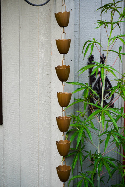 A vertical rain chain made of seven brown metal cup links hanging in front of a white wooden wall, with green plant leaves partially obscuring the right side.