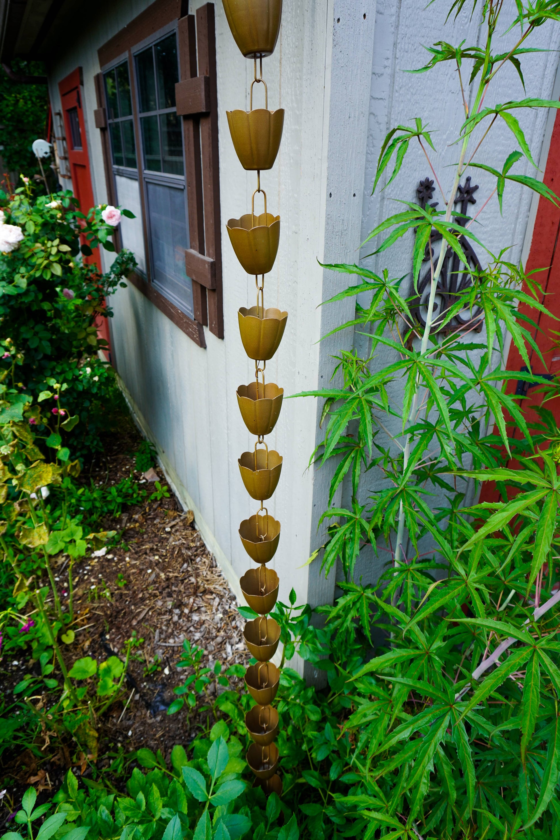 A vertical rain chain made of stacked metal pots hanging outdoors next to a house wall, surrounded by green plants and foliage.