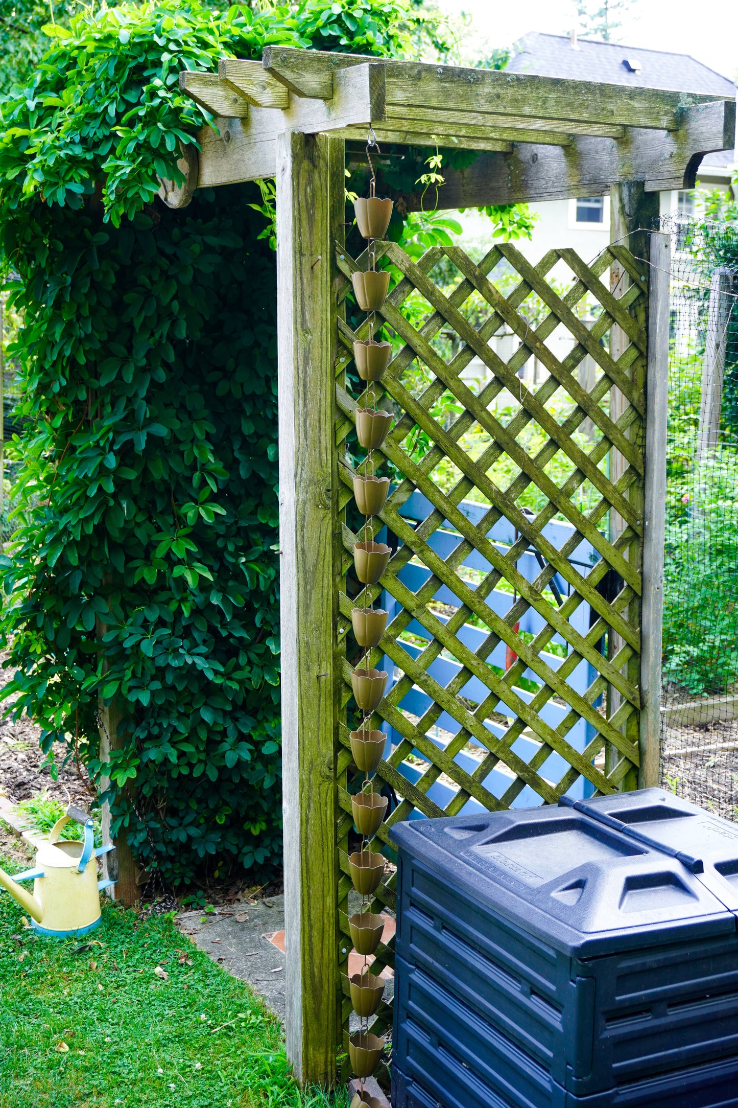 A wooden garden trellis arch with a lattice panel and hanging flower pots, shown in a backyard garden next to a black compost bin and green foliage.