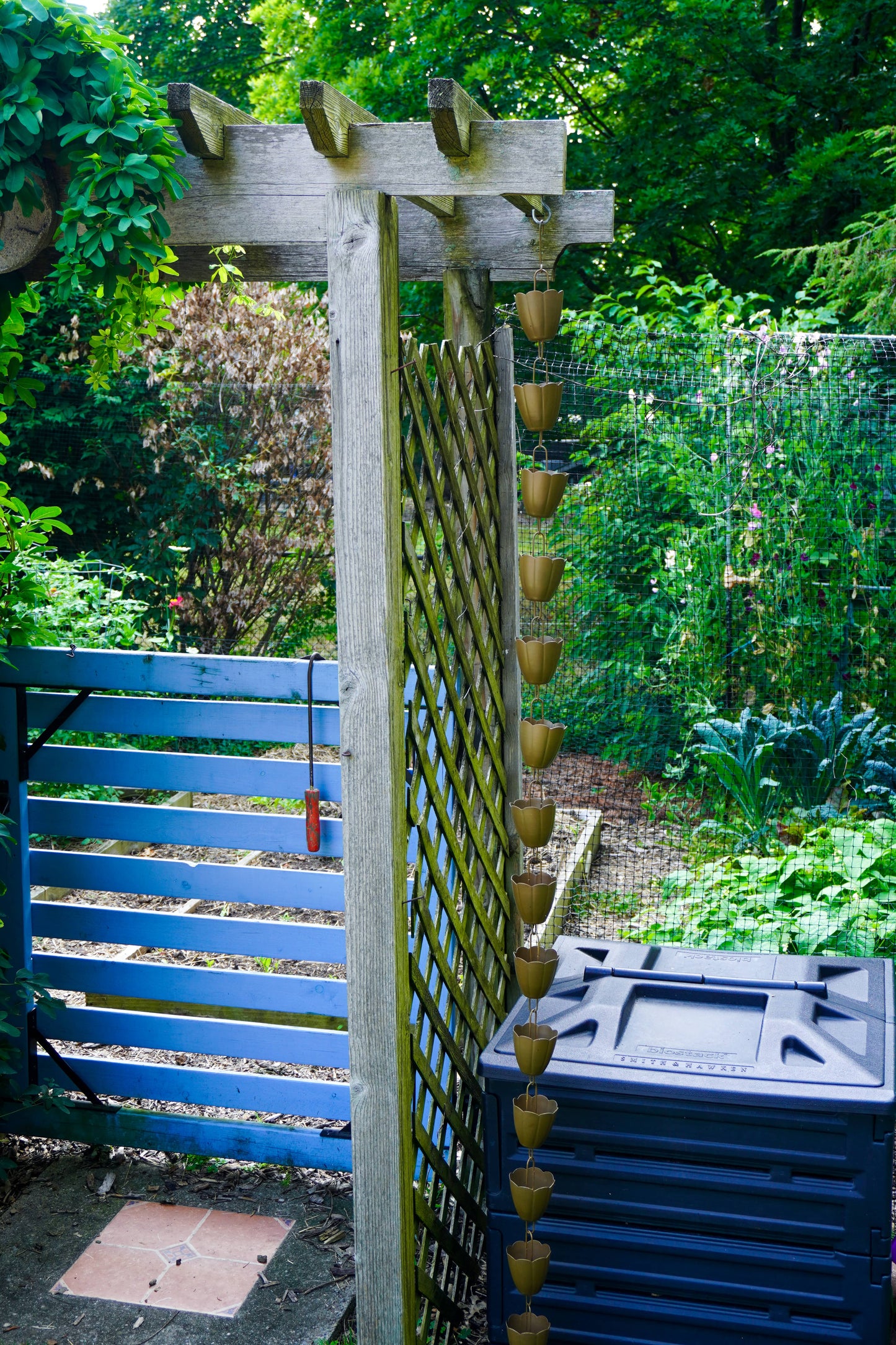 A wooden garden trellis with hanging rain gutters, set in a lush green backyard next to a blue wooden gate and a dark blue plastic storage bin.