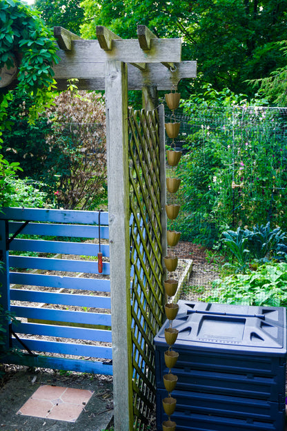 A wooden garden trellis with hanging rain gutters, set in a lush green backyard next to a blue wooden gate and a dark blue plastic storage bin.