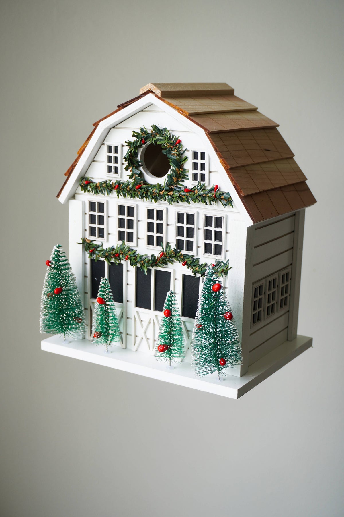 A decorative white barn-shaped birdhouse with a brown shingled roof, holiday garlands, a wreath, and small green Christmas trees with red ornaments, shown against a plain light gray background.