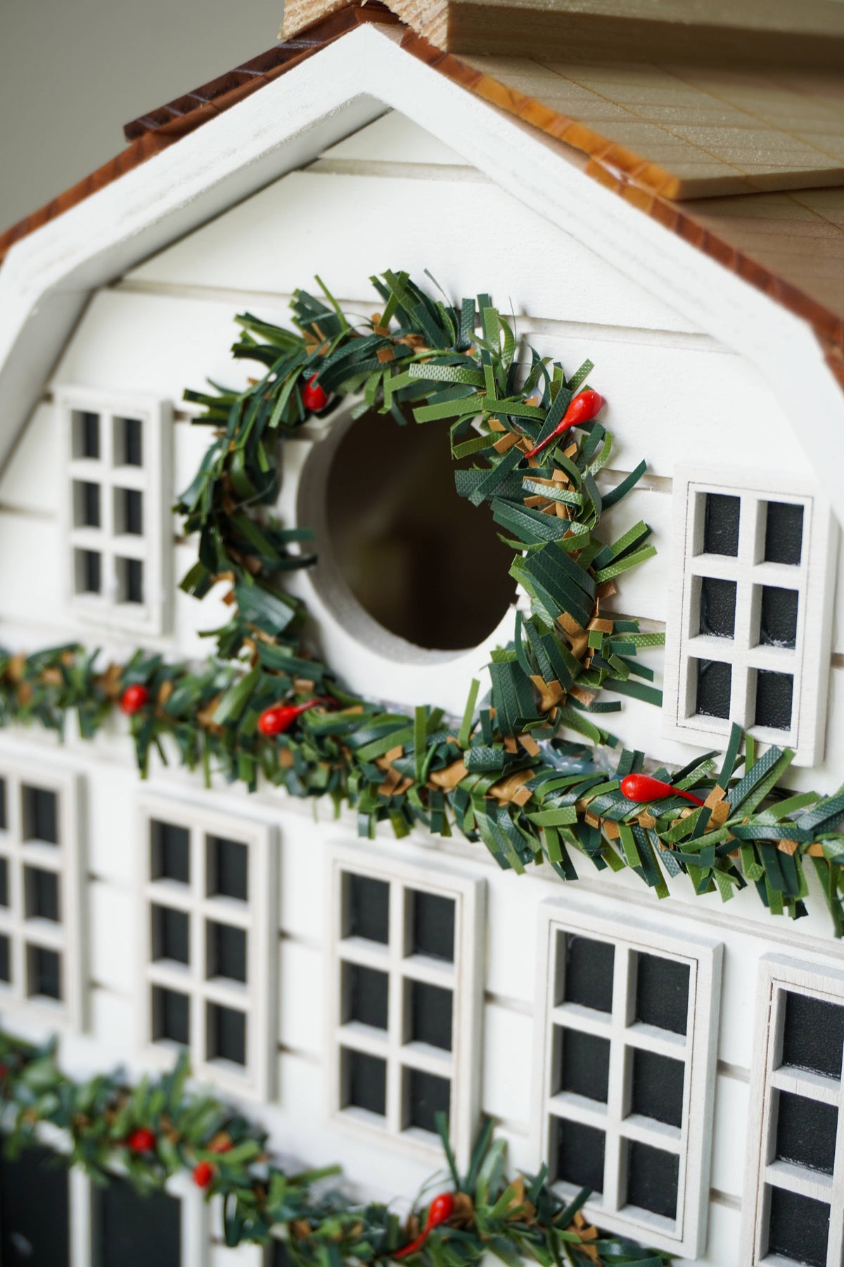 A close-up of a white holiday house ornament decorated with green pine garlands, small red berries, and a circular window opening.