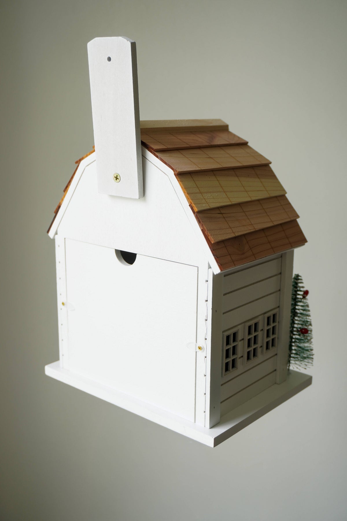 A white wooden birdhouse shaped like a cottage with a brown shingled roof, a small chimney, and a tiny green Christmas tree on the side, shown against a plain light gray background.