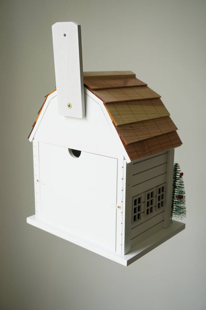 A white wooden birdhouse shaped like a cottage with a brown shingled roof, a small chimney, and a tiny green Christmas tree on the side, shown against a plain light gray background.