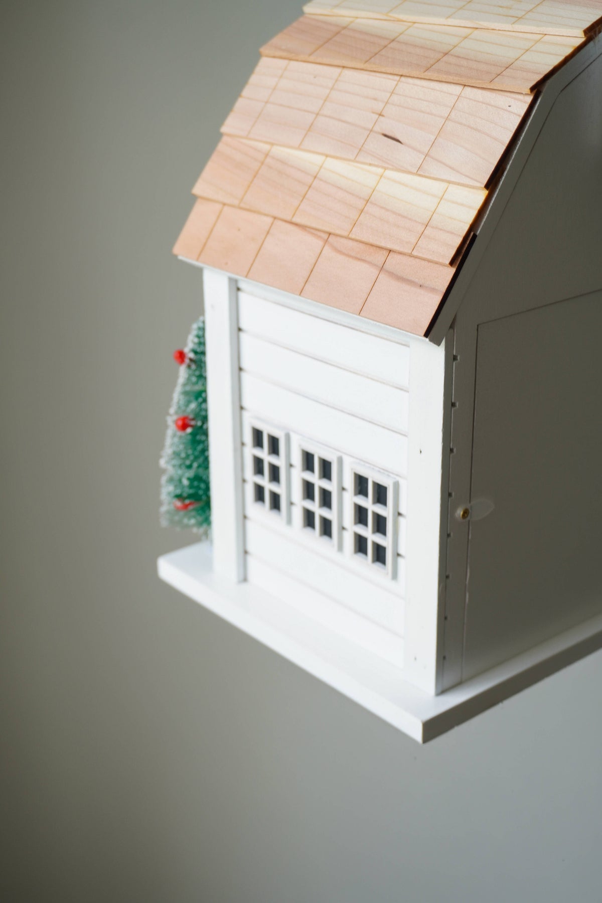A white wooden birdhouse with a light wood shingled roof, decorative window, and a small green Christmas tree ornament on the side, shown against a plain gray background.