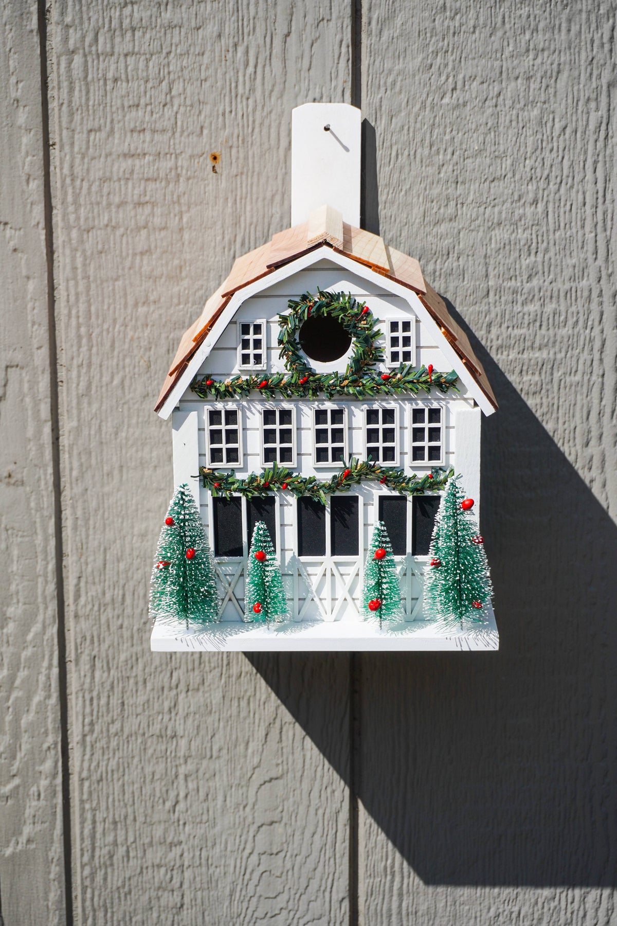 A white Christmas birdhouse shaped like a house with a brown roof, decorated with a green wreath, garland with red berries, and three small green Christmas trees, mounted against a gray wood-paneled wall.