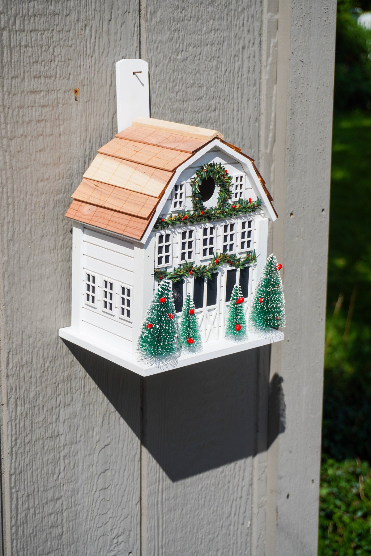 A white Christmas birdhouse shaped like a house with a brown shingled roof, green wreath, and small decorated trees, mounted on a gray wooden post outdoors.