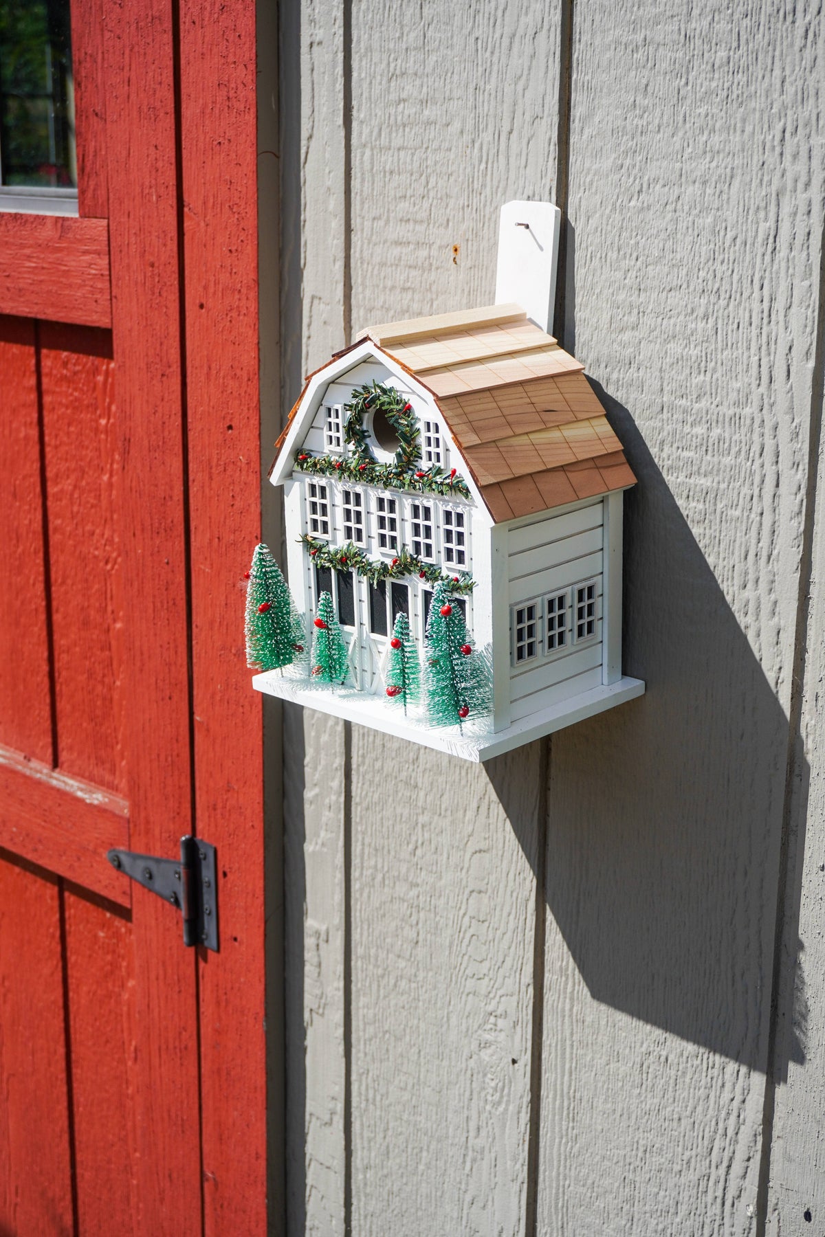 A decorative birdhouse shaped like a white farmhouse with a brown shingled roof, green wreath, and miniature Christmas trees, mounted on a gray wood wall next to a red door.