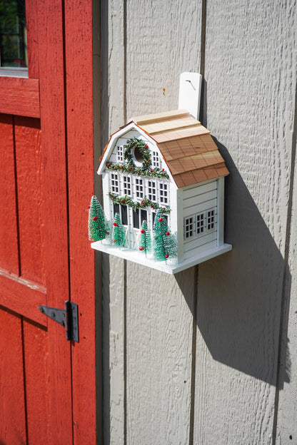 A decorative birdhouse shaped like a white farmhouse with a brown shingled roof, green wreath, and miniature Christmas trees, mounted on a gray wood wall next to a red door.
