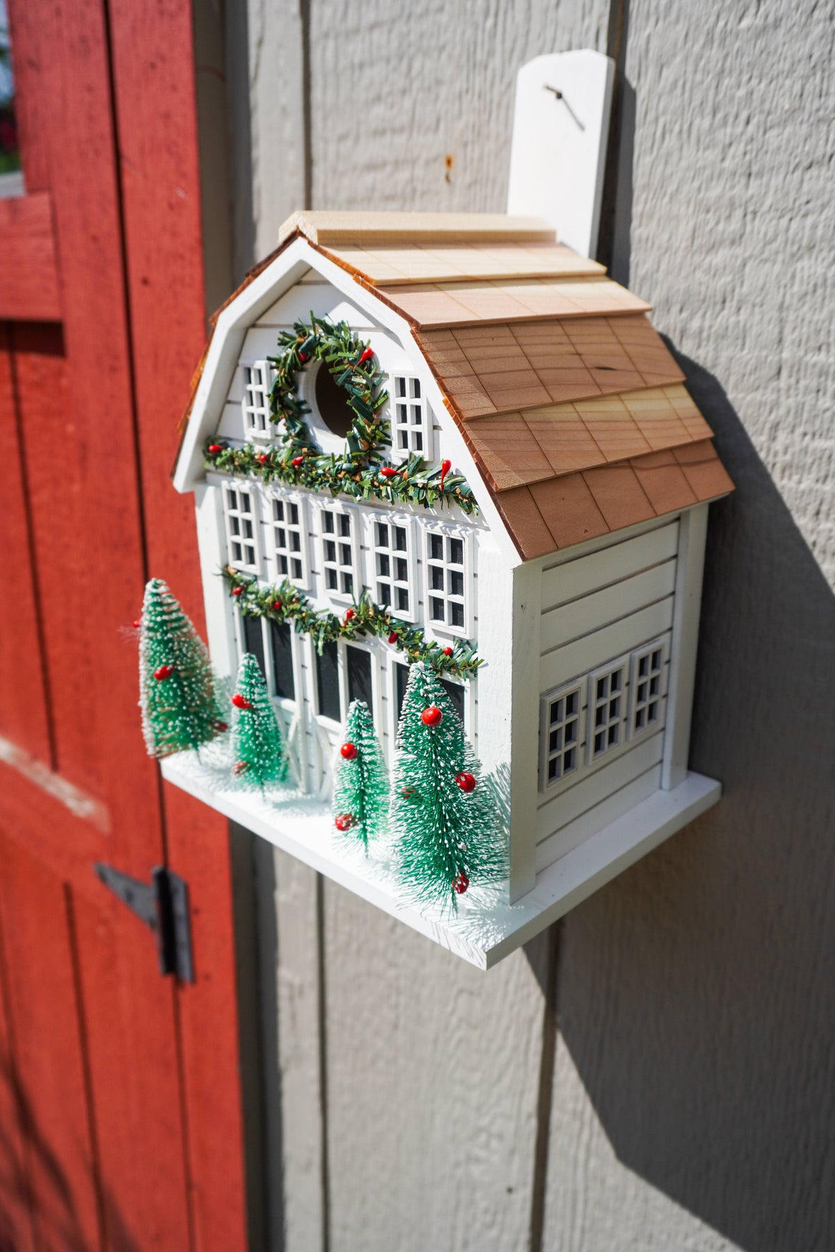 A decorative white birdhouse shaped like a small house with a brown shingled roof, holiday wreath, garlands, and four small green Christmas trees, mounted against a gray wall next to a red door.
