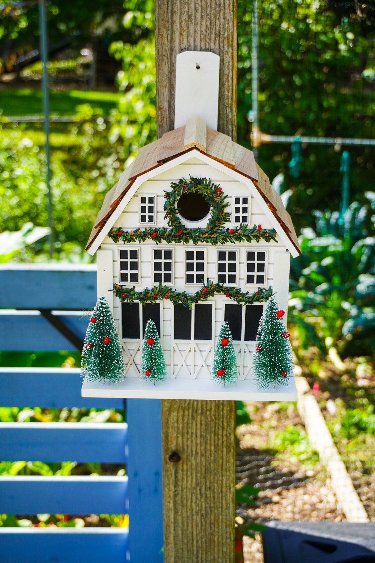 A white Christmas-themed house-shaped birdhouse ornament hanging from a wooden post in a garden. The ornament features a brown roof, black window panes, a green wreath with red berries on the front, and three small green Christmas trees at the base.