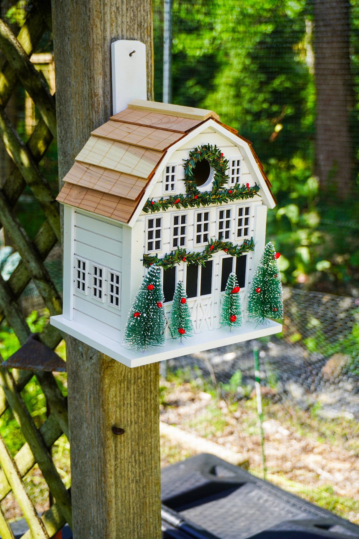 A decorative white holiday birdhouse shaped like a small house with a brown shingled roof, green garland details, a wreath on the front, and three small Christmas trees on the front porch, shown hanging outdoors on a wooden post with a garden background.