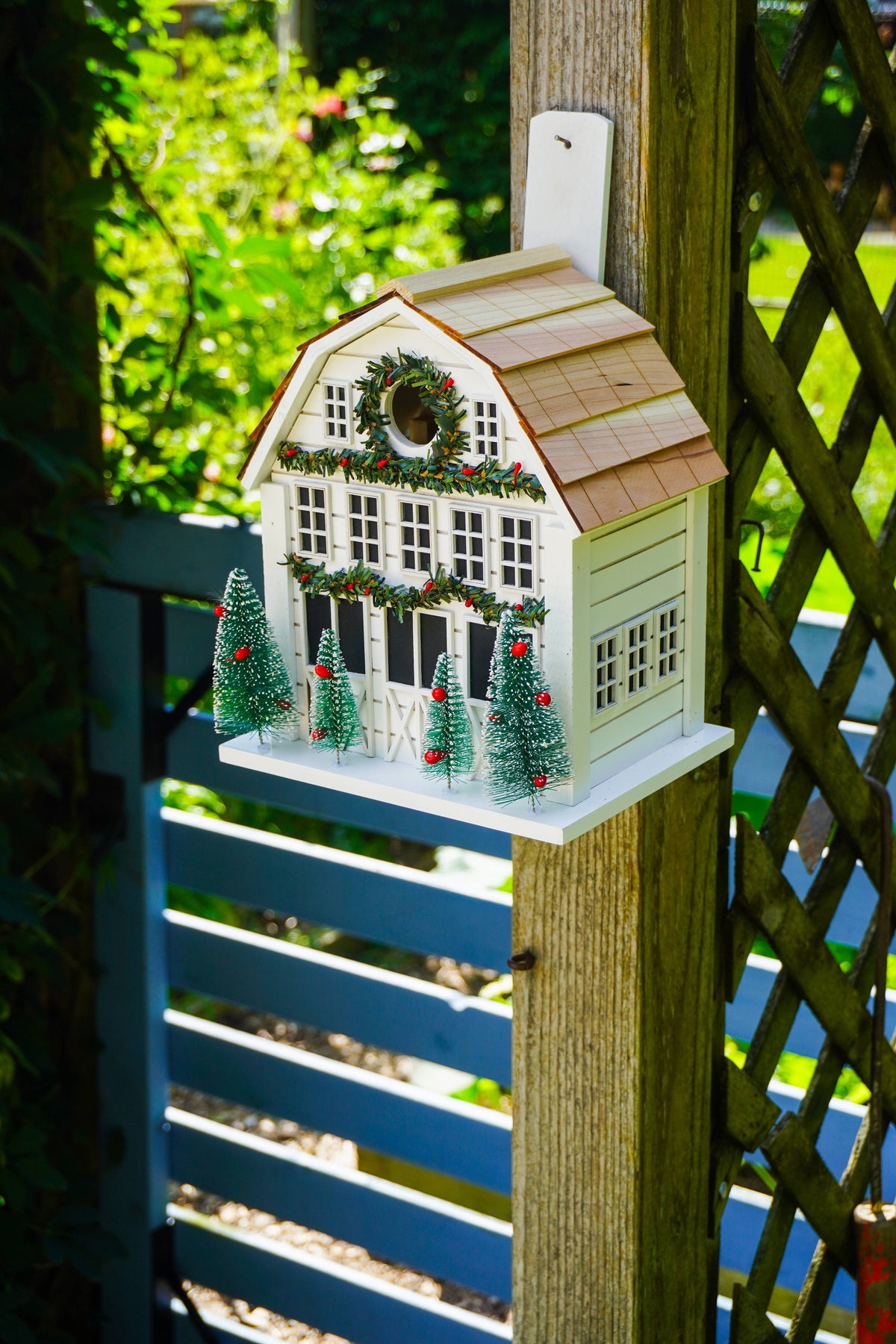 A white Christmas house ornament or decorative birdhouse hanging on a wooden post in a garden setting, with small green trees and red berries around it.