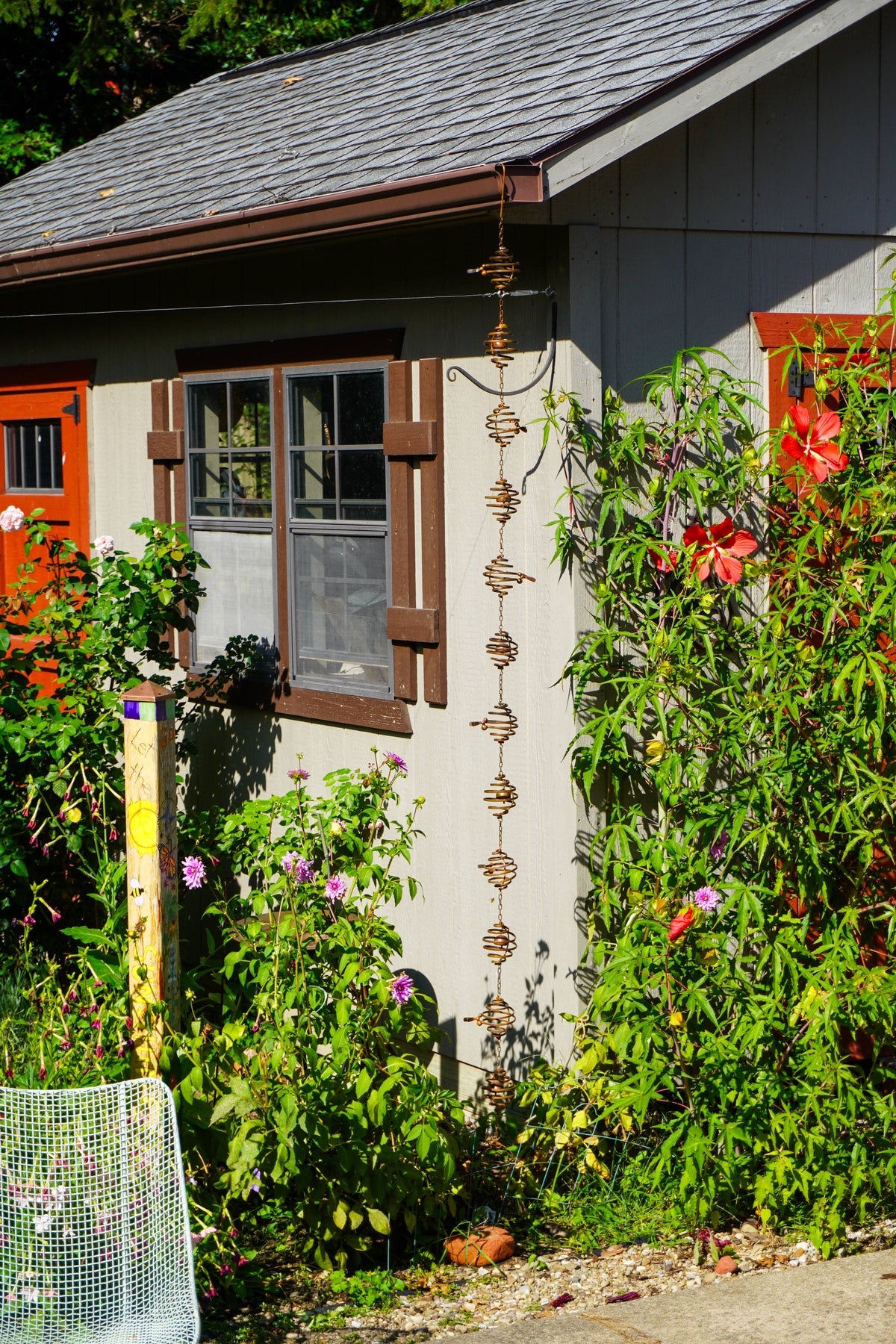 A vertical metal garden rain chain hangs from the gutter of a gray house, surrounded by lush green garden plants, red flowers, and orange shutters.