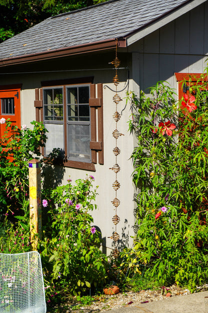 A vertical metal garden rain chain hangs from the gutter of a gray house, surrounded by lush green garden plants, red flowers, and orange shutters.