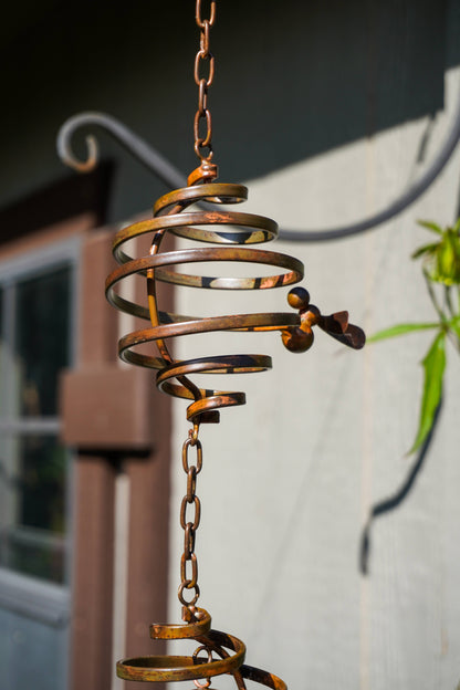 A rustic metal spiral garden wind spinner hanging from a chain outdoors against a light-colored wall with a blurred window and plant in the background.