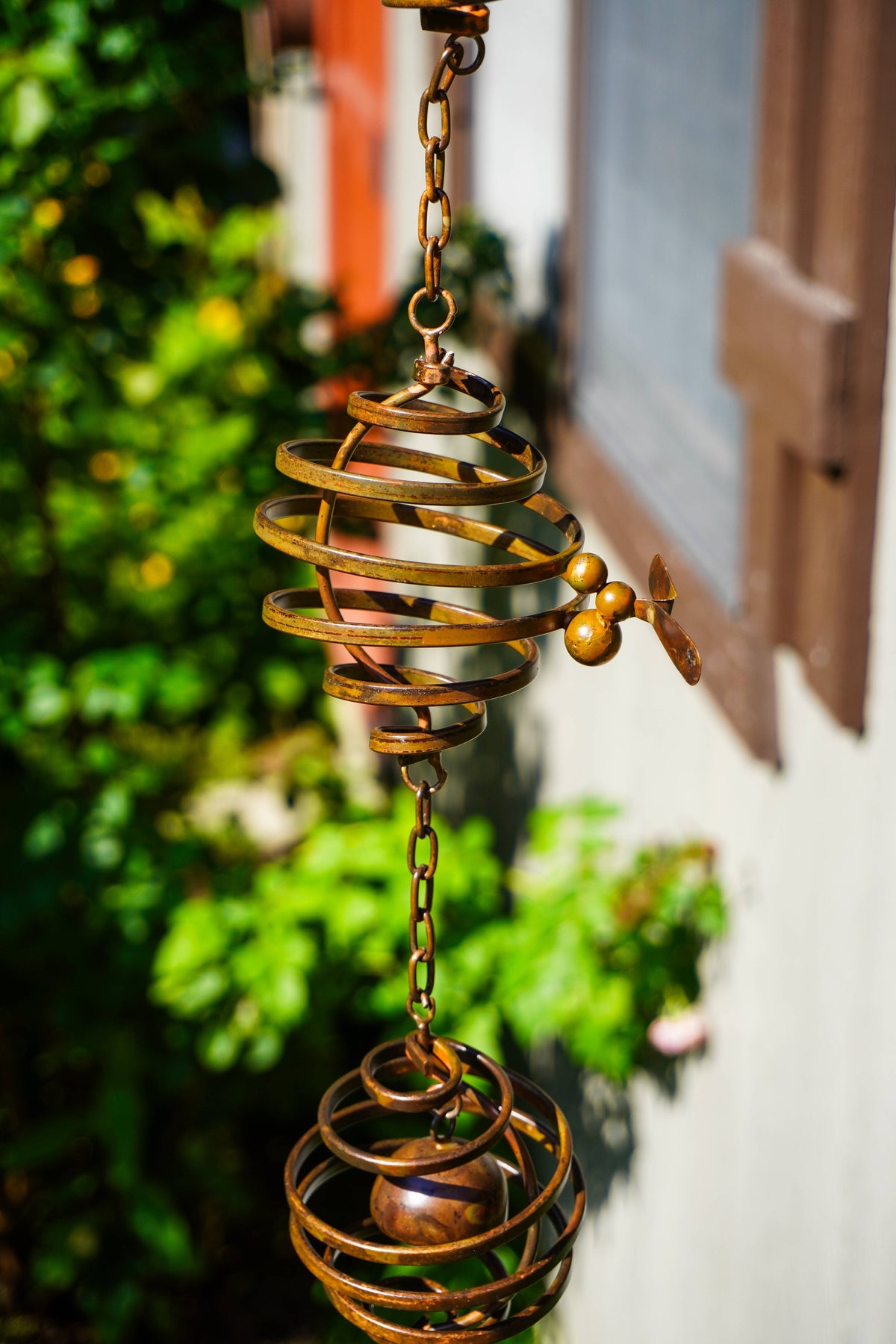 A brass wind spinner hanging from a chain outdoors, featuring two spherical wire cages and a small winged wheel, with blurred green foliage and a building wall in the background.