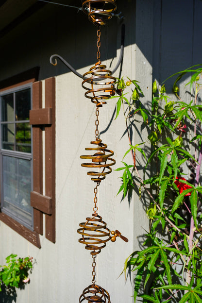 A rusted metal spiral wind spinner hanging from a bracket against a house exterior wall, with green plants and a window visible in the background.