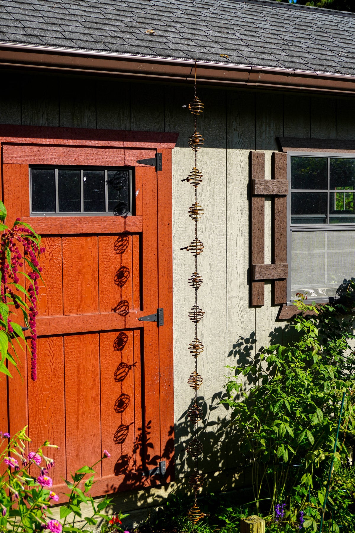 A vertical metal garden rain chainwith a beehive design hangs in front of a rustic red barn door and a light gray house wall, surrounded by green garden foliage and pink flowers.