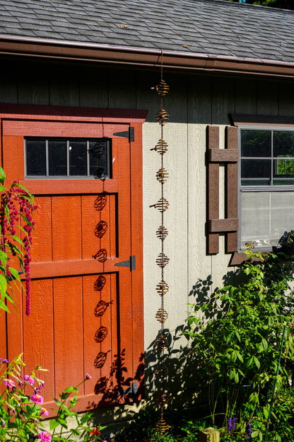 A vertical metal garden rain chainwith a beehive design hangs in front of a rustic red barn door and a light gray house wall, surrounded by green garden foliage and pink flowers.