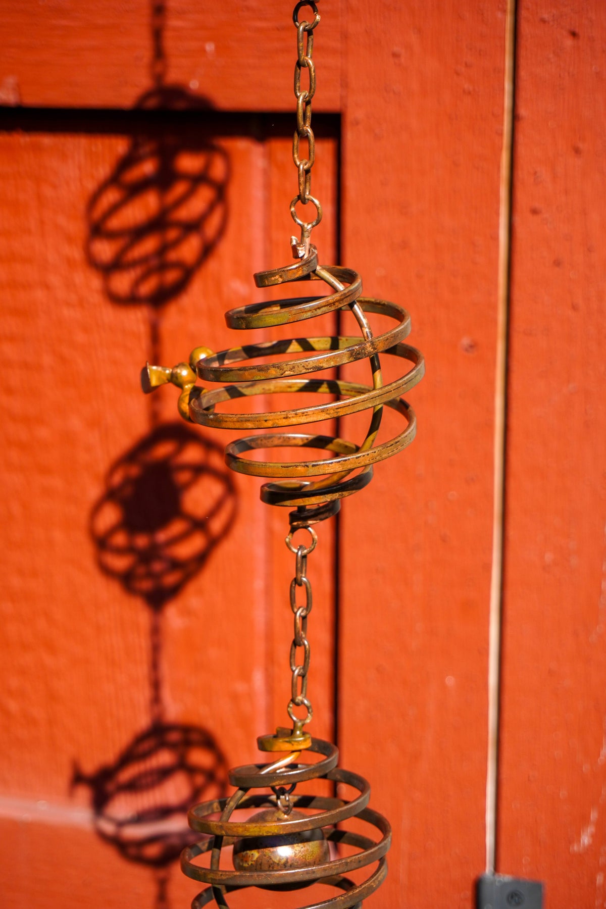 A brass hanging spiral ornament with a chain, photographed against a bright red-orange wooden door with strong shadows cast on the background.