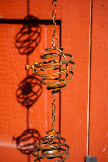 A brass hanging spiral ornament with a chain, photographed against a bright red-orange wooden door with strong shadows cast on the background.