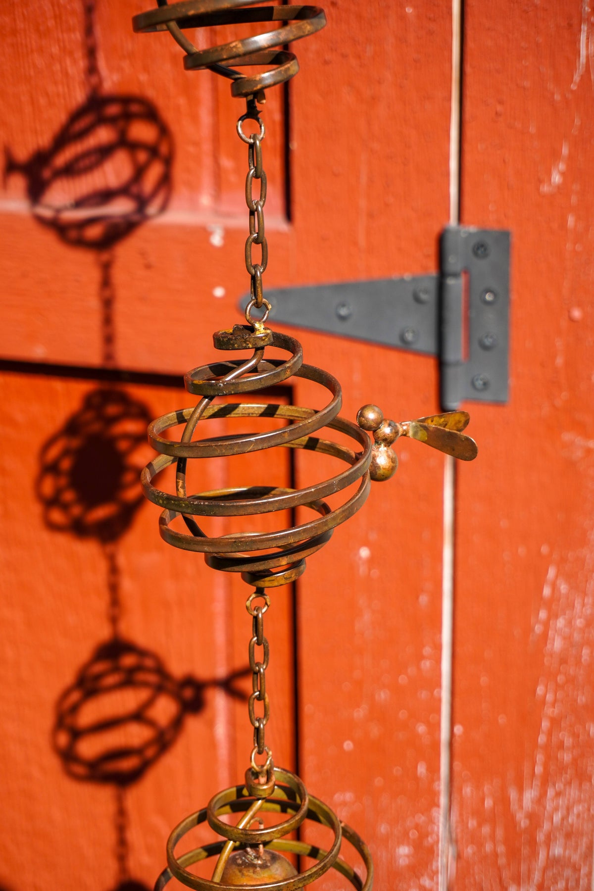 A brass wind spinner with spiral wire spheres and a small leaf blade, hanging in front of a textured orange-red wooden door.