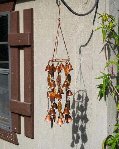 A copper wind chime with multiple small bells and leaf-shaped metal pieces hangs from a curved black metal hook on a light gray wooden wall. A shuttered window is visible on the left, and green leaves cast shadows on the wall to the right.
