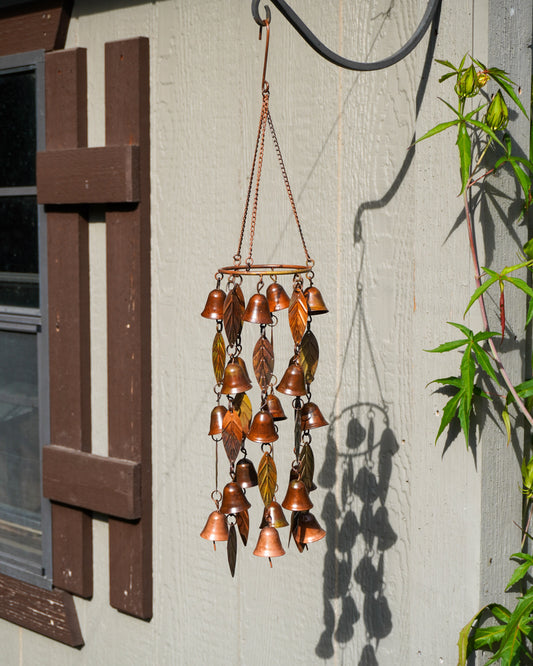 A copper wind chime with multiple small bells and leaf-shaped metal pieces hangs from a curved black metal hook on a light gray wooden wall. A shuttered window is visible on the left, and green leaves cast shadows on the wall to the right.