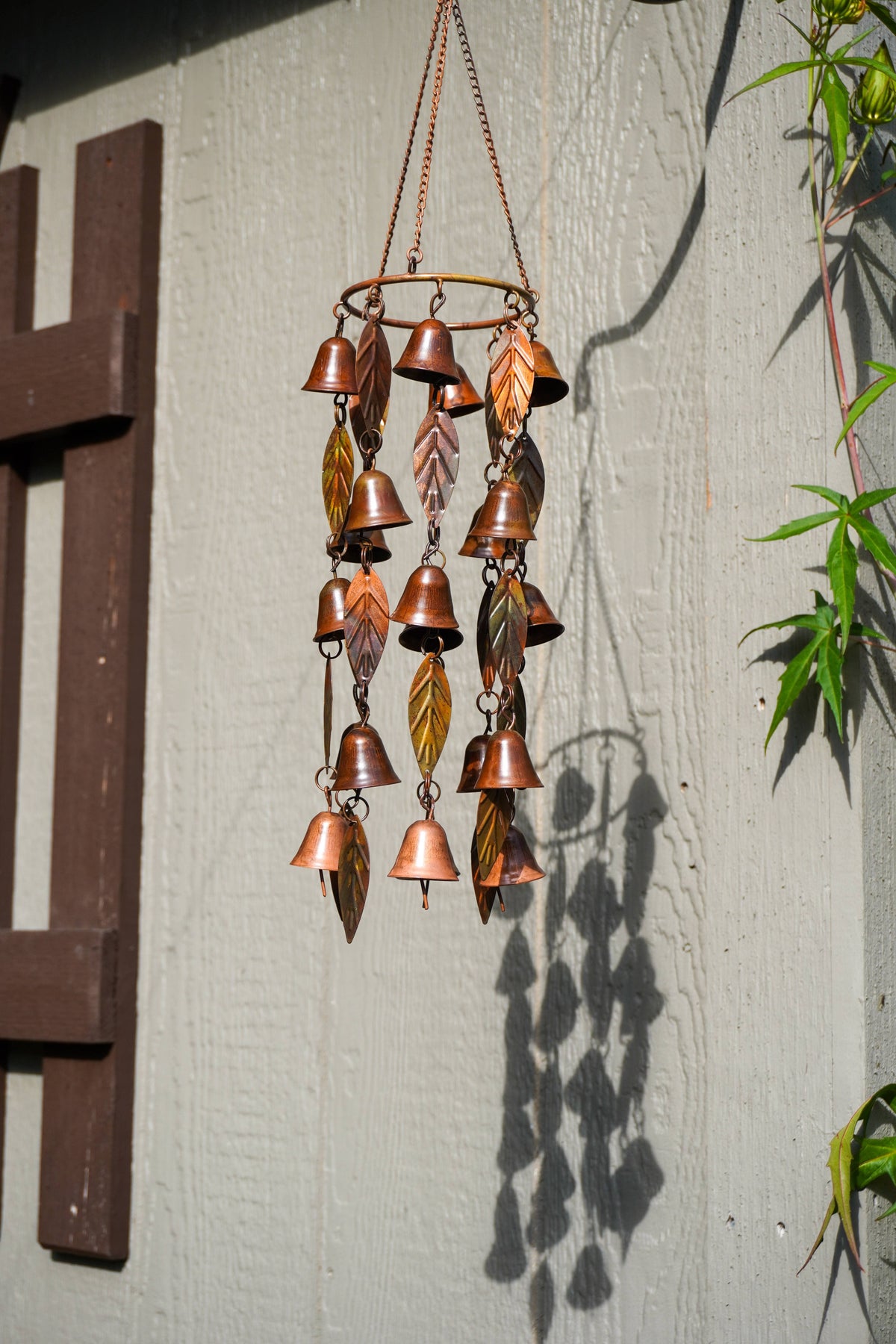 A copper wind chime hanging outdoors against a light gray wall, featuring multiple small bell-shaped and leaf-shaped metal pieces in a warm copper tone.