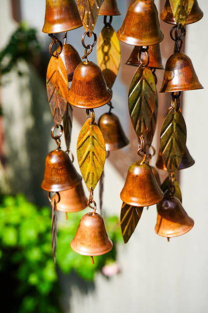 Copper wind chimes shaped like small bells and leaves, hanging in strands against a softly blurred outdoor background.