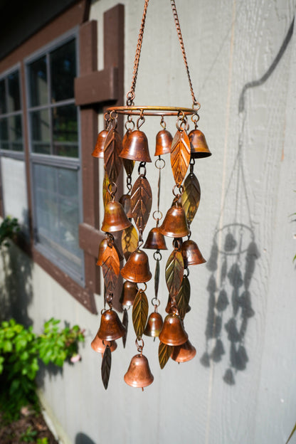 A copper wind chime hanging outdoors against a light gray wall, featuring multiple small bell-shaped and leaf-shaped metal pieces in a warm copper tone.