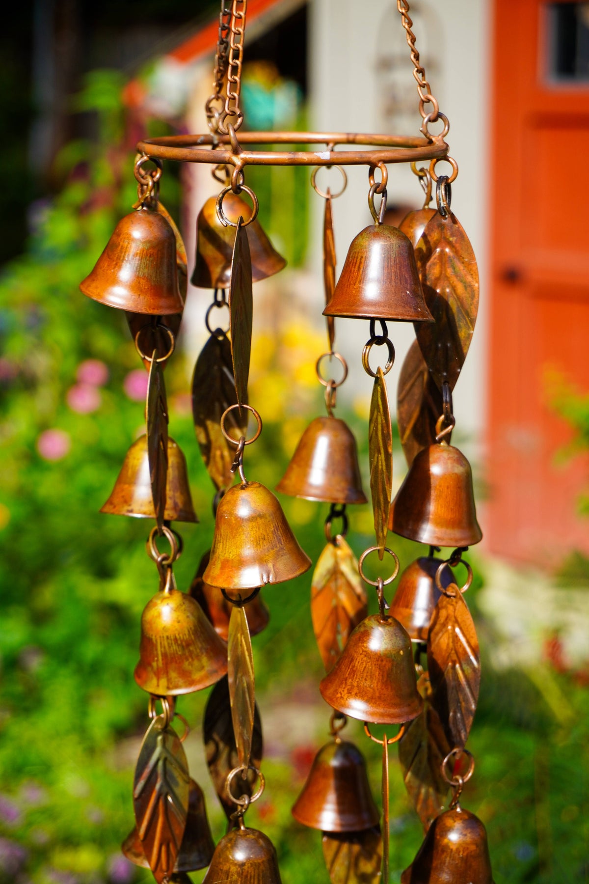 A hanging wind chime made of multiple small metallic bells and leaf-shaped pieces, photographed outdoors against a blurred garden and red-orange building background.